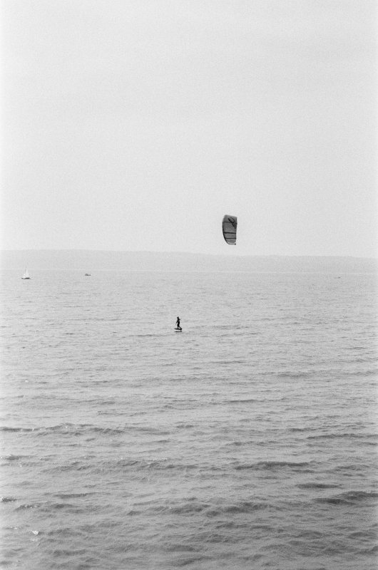 A blown out and somewhat soft photo of the dee estury with wales in the background. In the middle of this frame is a small but contrsting pair of shapes - a surfer on a hydrofoil board and above them a parachutte shaped kite that is pulling them along.