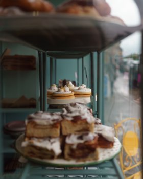 A view of a plate of iced biscuits sat on some shelves in a shop window, surrounded by other out of focus cakes.