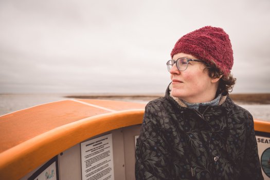 A photo of Laura sat in the front of an orange boat. Laura is in warm weather gear and looking out to sea.