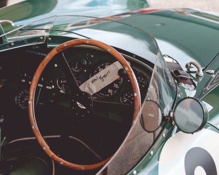 The dashboard of a vintage Aston Martin race car, over the main dial of which has been taped some masking tape with "New Tyres!" hand written on it.