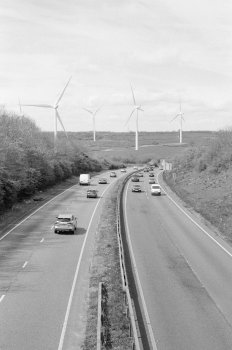 A photo looking from a bridge over a dual carridgeway road with cars on it. The road slowly sweeps to the right, and then straight ahead are a row of four wind turbines, standing tall over the landscape.