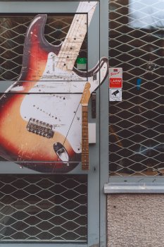 A photo of a door to a guitar shop, part covered with a large sticker of a fender stratocaster. The door handle is an entire next from a Telecaster style guitar.