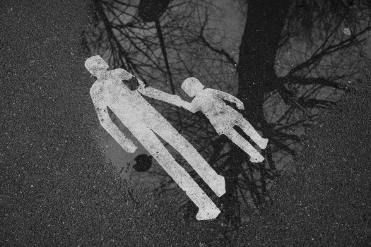 A photo of some pavement, on which is painted a sign of an adult walking hand in hand with a child. The sign is in a puddle which is showing the reflection of a bare tree. The sign is solid white paint, but has a slight sense of depth to it unlike those found in the UK.
