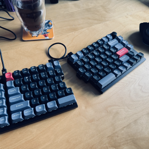 A photo of a split mechanical keyboard, in black with a mix of black, blue, and red keycaps. It sits on a desk, next to the keyboard is a trackball and a cup of coffee.