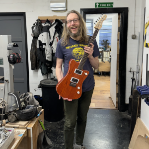 A photo of me in a workshop holding an orange electric guitar, smiling. The guitar is made from wood stained a bright orange with dark grain patterns, and has a maple neck with a rosewood fretboard.