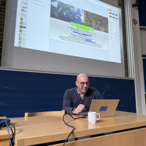 A photo of Anil sat at a desk at the front of a lecture theatre with a laptop, and behind him is a projection of a title slide of a talk `Programming for the planet` with both his name and mine as presenters.